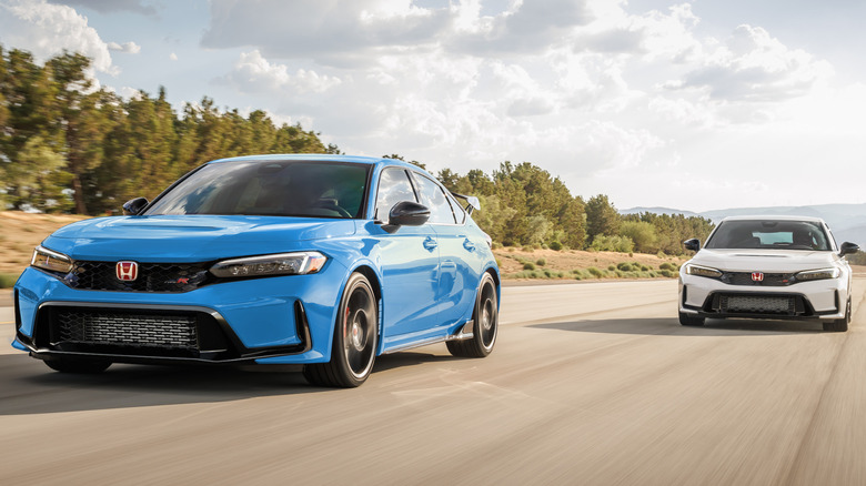 Front three-quarters shot of a blue Honda Civic Type R driving straight on a track in front of a white Civic Type R behind it as they pass green trees and a cloudy sky