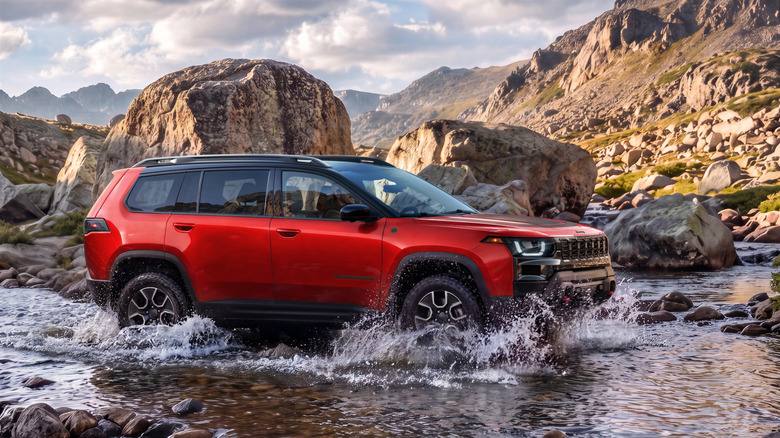 Side view of a red Cherokee Trailhawk driving through water in front of rocks and a scenic mountain backdrop