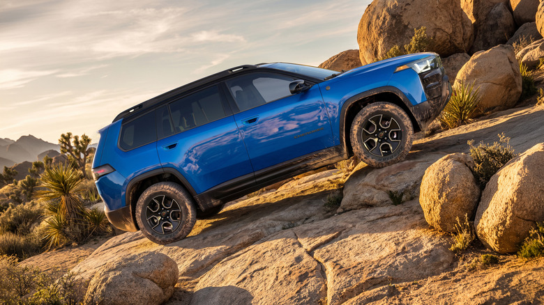 Side view of a blue Cherokee Trailhawk driving up a rock incline in front of Joshua Trees and more rocks