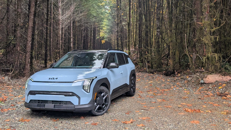 a Frost Blue 2027 Kia EV3 parked in front of some old growth pine trees in the pacific northwest