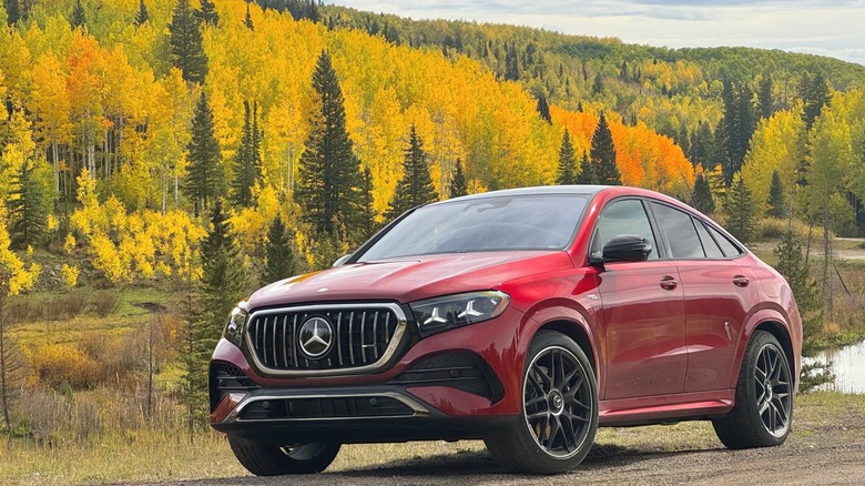 Front three-quarters shot of a red GLE53 AMG Coupe parked on dirt in front of a mountain covered in bright yellow trees