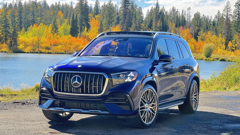 Front three-quarters shot of the dark blue GLS63 press car parked on asphalt in front of a lake that's surrounded by bright yellow and green trees and a cloudy sky