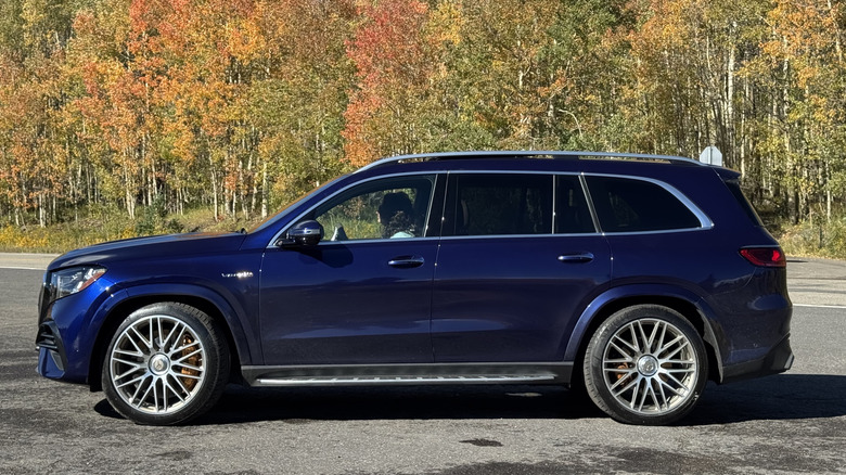 Side view of the dark blue GLS parked on asphalt in front of yellow, green, and red trees