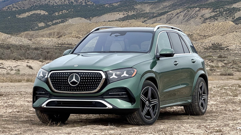 Front three-quarters shot of a light green 2027 GLE500e parked on dry dirt in front of a shrub-covered mountain