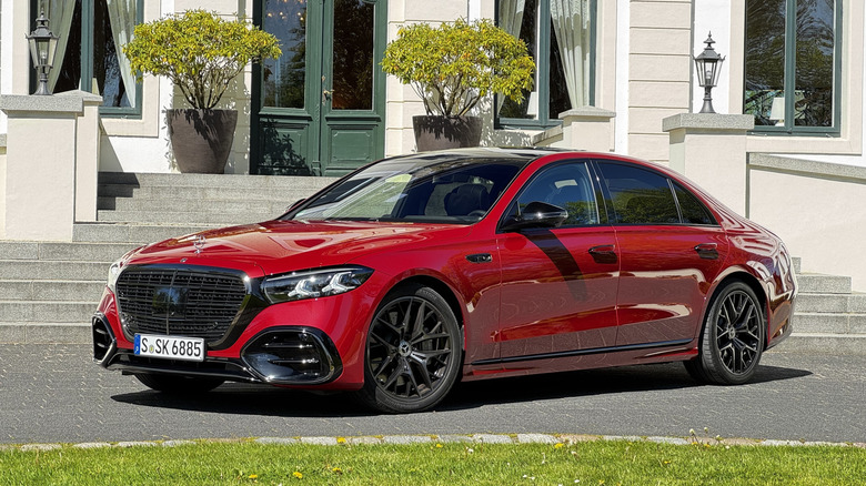 Front 3/4 view of a deep red S580 parked behind grass in front of a white building with a porch and two trees next to the door