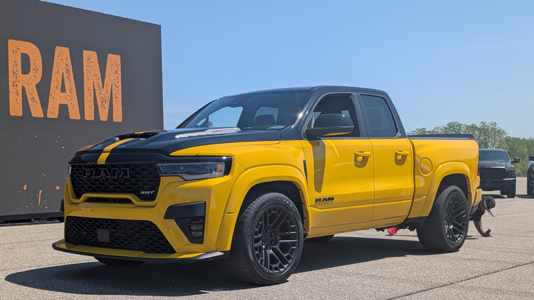 The front 3/4ths view of a bright yellow Ram 1500 Rumble Bee SRT parked on the Chelsea Proving Grounds racetrack with the word "RAM" in big orange letters in the background