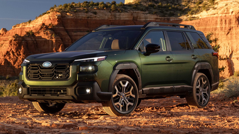 front three-quarters shot of a dark green 2026 Outback parked on dirt in front of a red cliff at sunset