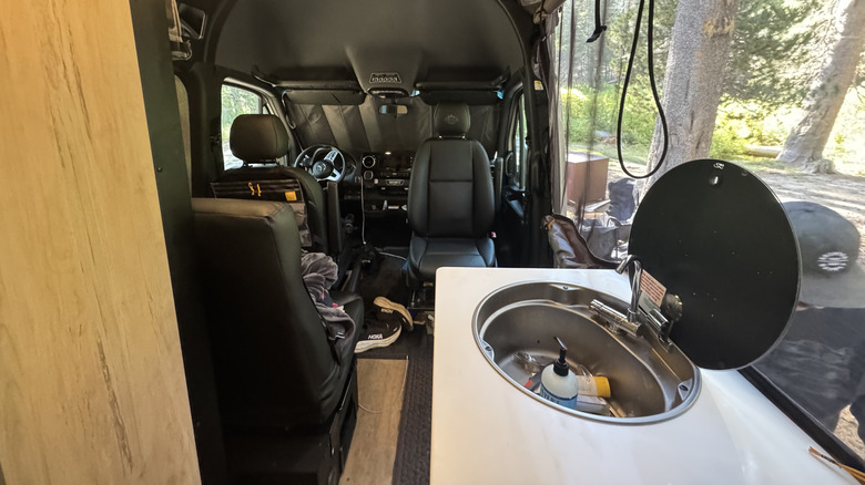 A look forward from the bed showing the open sink, rotated passenger seat, front windshield cover, and trees outside