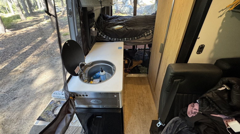 A photo looking back at the interior of the van, showing the sink and the bed deployed with a  view of dirt and roxks out the doors
