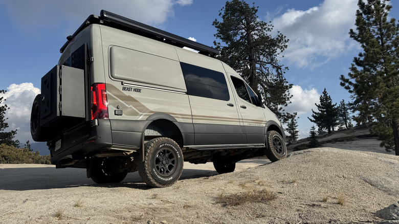 A rear three quarters shot of the van with its front right wheel propped up on a boulder in front of a blue sky and trees