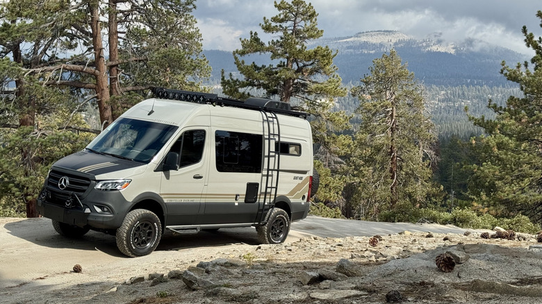A front three quarters shot of the van parked on a narrow rocky trail with trees and a view of a mountain in the background