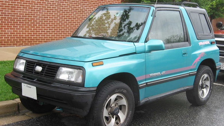 A teal green Geo Tracker Convertible parked on the street by a red brick wall.