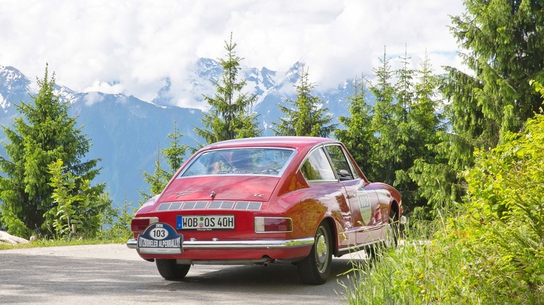 A red Karmann Ghia TC 145 parked on a mountain road with trees, fog, and snow-capped mountains in the distance