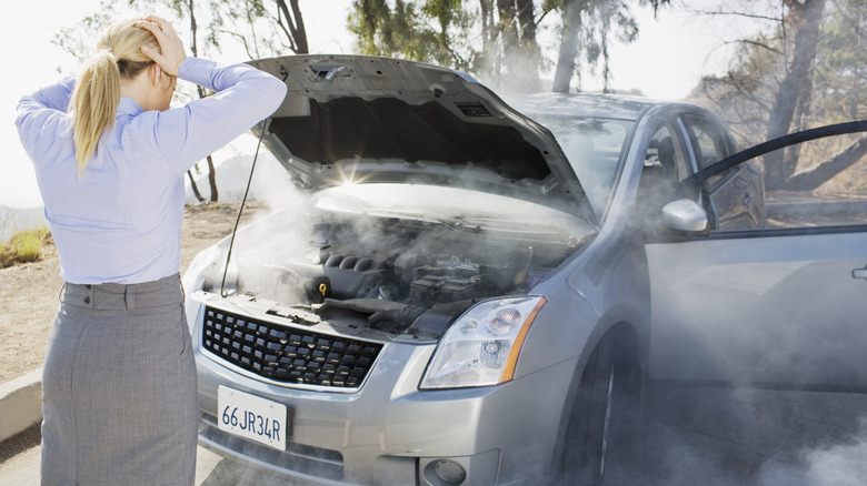 A person is standing in front of an overheating gray car with its hood up, looking distressed.
