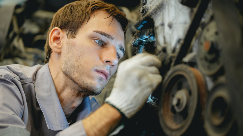 A young man wearing white gloves inspects a junkyard engine