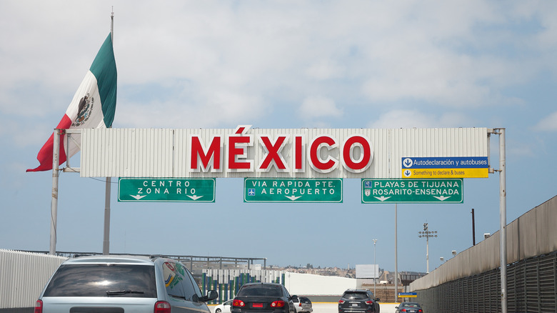 The border car entrance point to Baja, Mexico from the USA with cars on the road and a Mexican flag and sign.