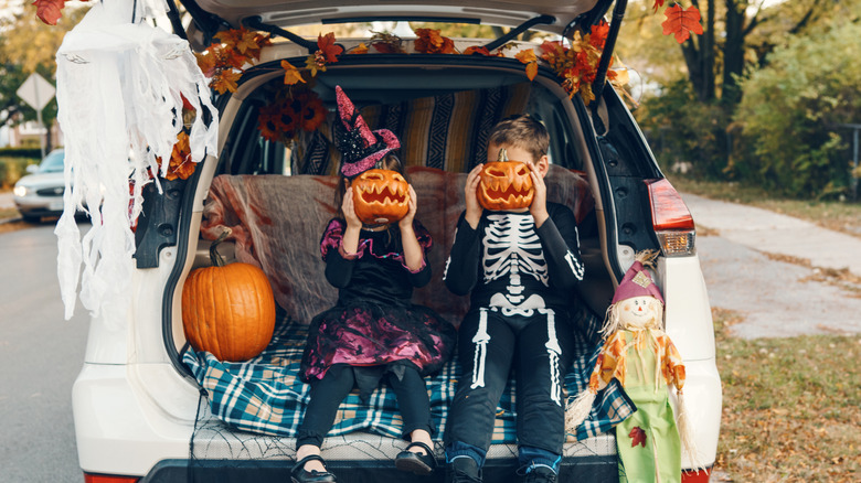 Two kids holding pumpkins to their faces sitting on the trunkbed with Halloween decorations around them.