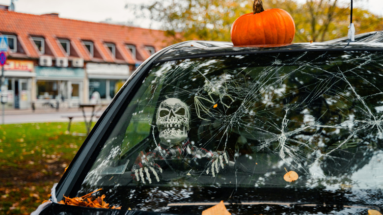 A skeleton placed in the passenger's seat of a car with cobwebs and a pumpkin on the roof.