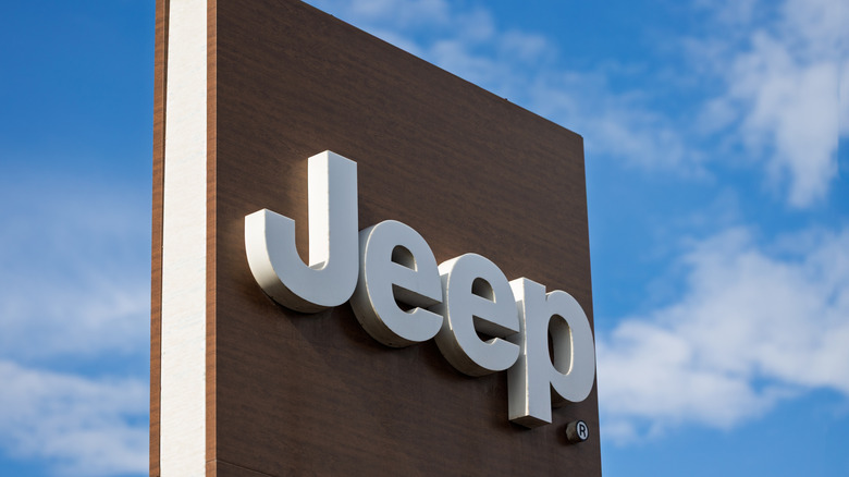A silver Jeep logo mounted on a wooden-finished pillar with the sky in the background on a sunny day.