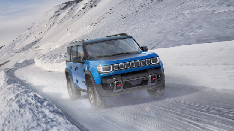 A metallic blue 2026 Jeep Recon driving along a snow-covered mountain road.