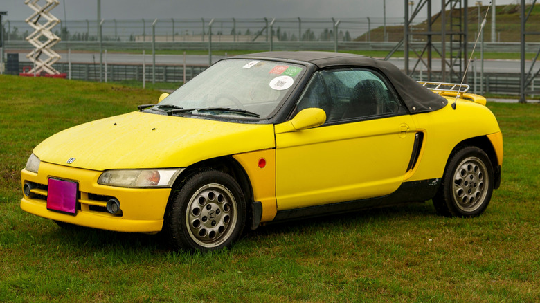 A yellow Honda Beat kei car parked on grass near a track.