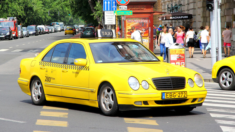 A yellow Mercedes W210 taxi being driven on busy European city roads.