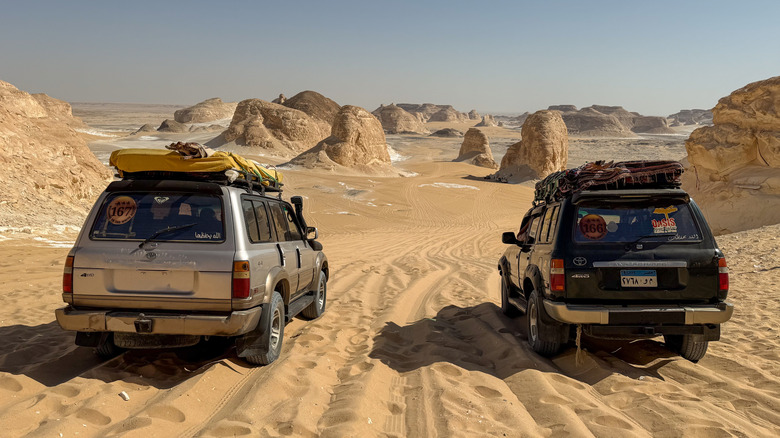 Two old Toyota Land Cruiser models parked on desert sand with desert hills in the background.