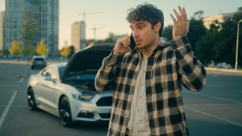 A frustrated man on the phone standing in front of a silver Mustang with its hood open in the city.