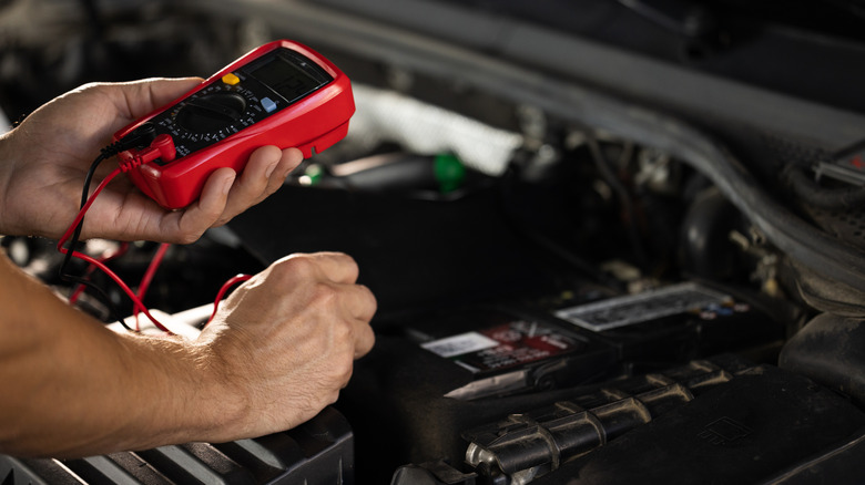 A person using a red multimeter to test a car's voltage output within the engine bay.
