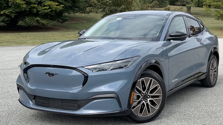 A blue-gray 2024 Ford Mustang Mach-E sits parked in an open lot with trees in the background