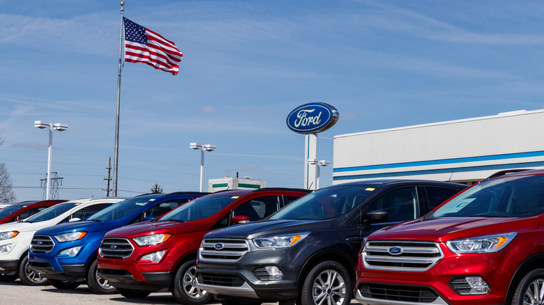 A Ford dealership sits below a blue sky as an American flag waves.