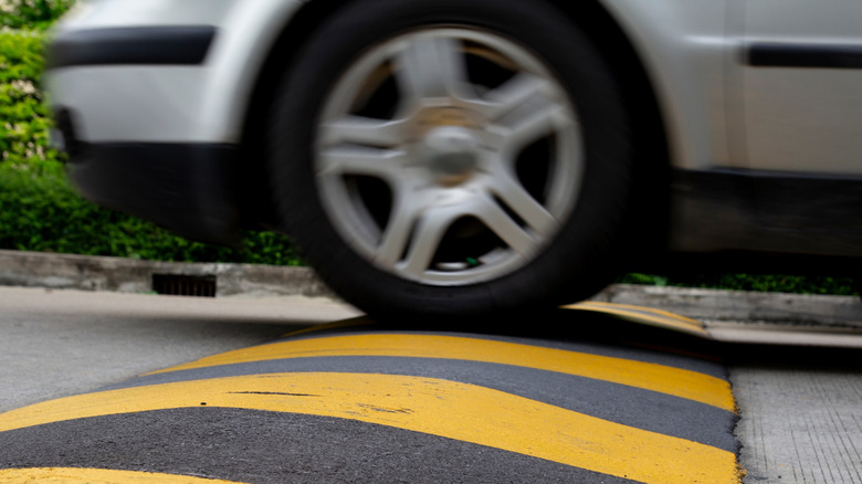A close-up view of a silver VW Golf Mark IV going over a black-and-yellow speed bump, with greenery in the background.