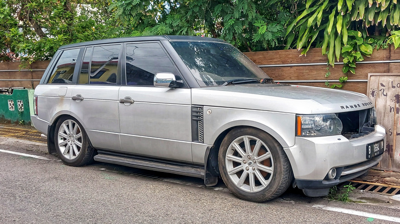 A silver old Range Rover luxury SUV parked on the roadside with collapsed air suspension, greenery in the background.