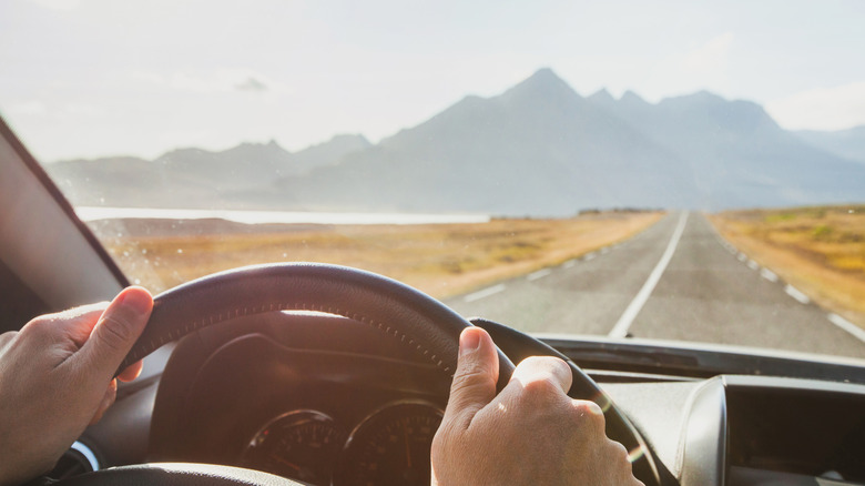 Close-up of hands on steering wheel, driving on straight road with mountains and golden fields under sunlight.