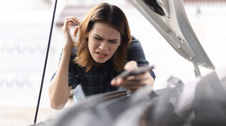 A frustrated woman standing over a car's open engine bay, listening with her right hand cupped next to her right ear.