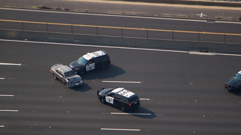 A grey saloon car with smashed front-end is hit on the side by a police SUV, with two other police cars nearby, on a main strip of highway