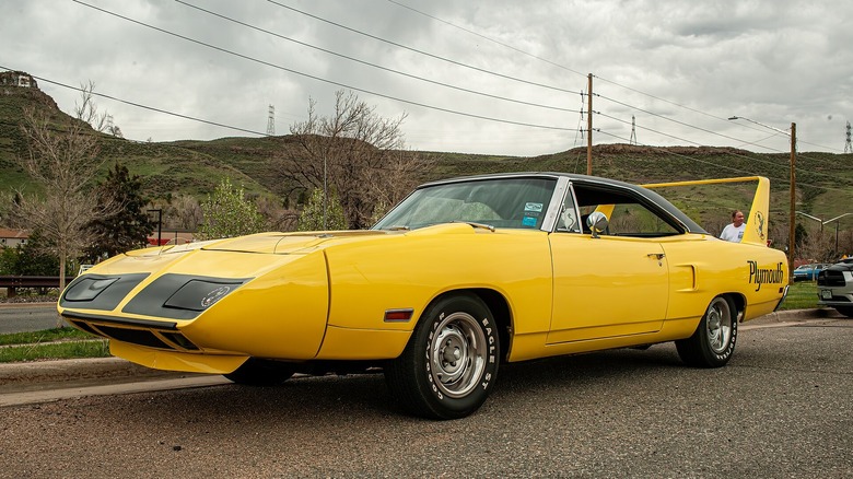 A bright yellow 1970 Plymouth Superbird showcasing its aerodynamic pointed nosecone, tall rear wing, and black hood graphics parked on asphalt.