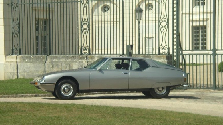 A sleek silver Citroën SM luxury grand tourer parked on gravel in front of tall ornamental iron palace gates.