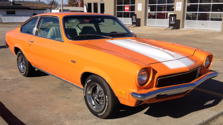 A bright orange Chevrolet Vega coupe with a white center hood stripe, chrome front bumper, and BFGoodrich white-letter tires parked outside a service garage.