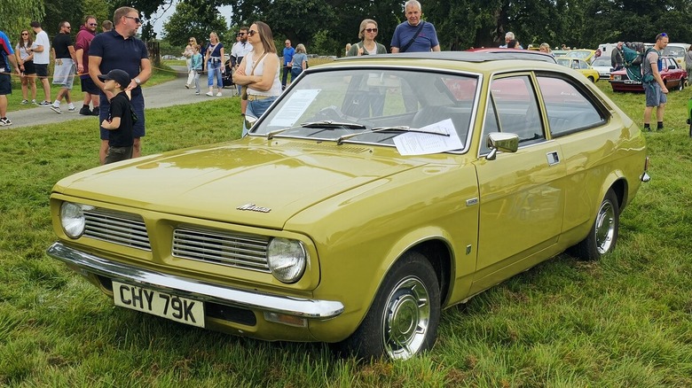 A mustard-yellow 1971 Morris Marina 1.3 SDL coupe with chrome hubcaps parked on grass at an outdoor car show.