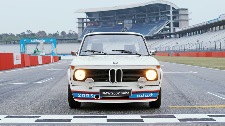 A white BMW 2002 Turbo with auxiliary driving lights and reverse 'turbo' front spoiler lettering sitting on a racetrack start-finish line.