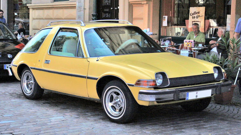 A bright yellow AMC Pacer hatchback with its distinctive bubble-glass greenhouse parked on a cobblestone street.