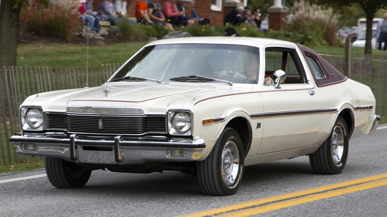 A white late-1970s Dodge Aspen coupe featuring a burgundy vinyl landau roof and heavy chrome bumpers driving on a road.