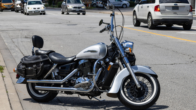 A black and white Honda Shadow parked on the street with numerous cars driving by in the background