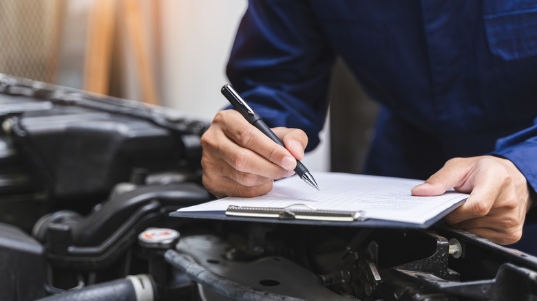 A mechanic holding a clipboard while inspecting a car's engine bay