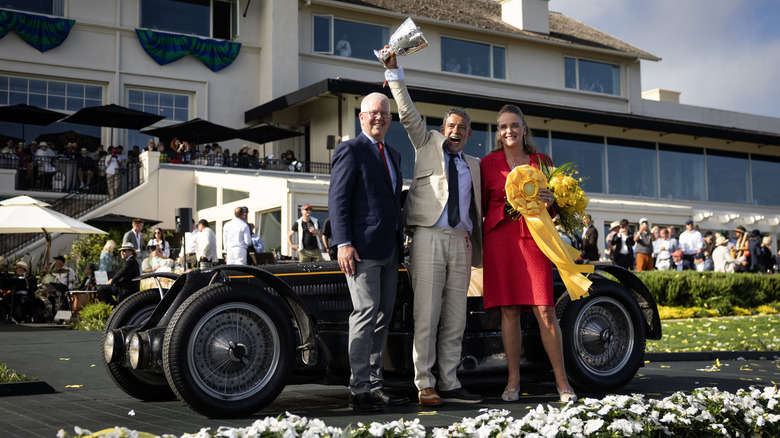 The 1934 Bugatti Type 59 Sports that won best in show at the 2024 Pebble Beach Concours d'Elegance sitting on a podium in front of a building with it's owner proudly hoisting the trophy in front of it.