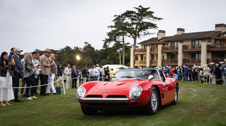 A red show car parked on grass at the Pebble Beach Concours D'Elegance event with people and a large building in the background