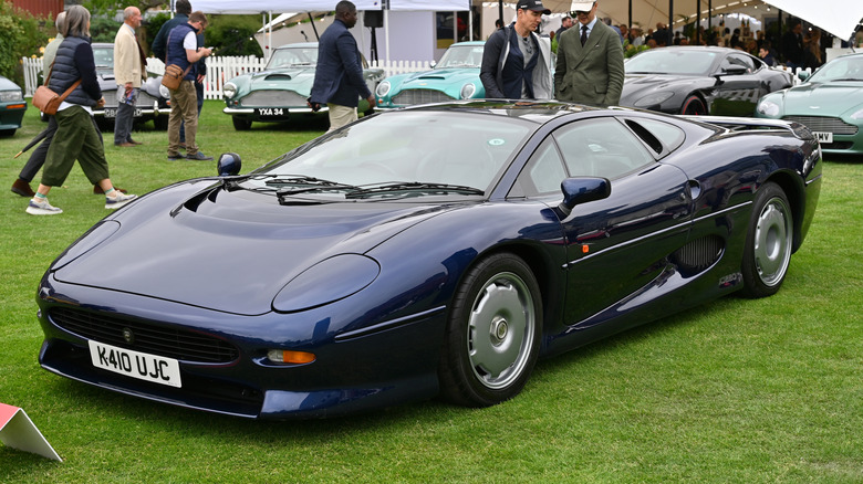 A dark blue Jaguar XJ220 parked on a lawn at a car show with other vehicles and people in the background