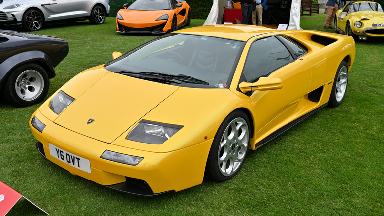 A yellow Lamborghini Diablo parked on a lawn at a car show with other luxury and vintage vehicles in the background