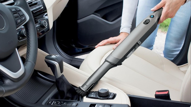 A Slate gray Shark WandVac with its brush tool attached is used to clean a car's center console.